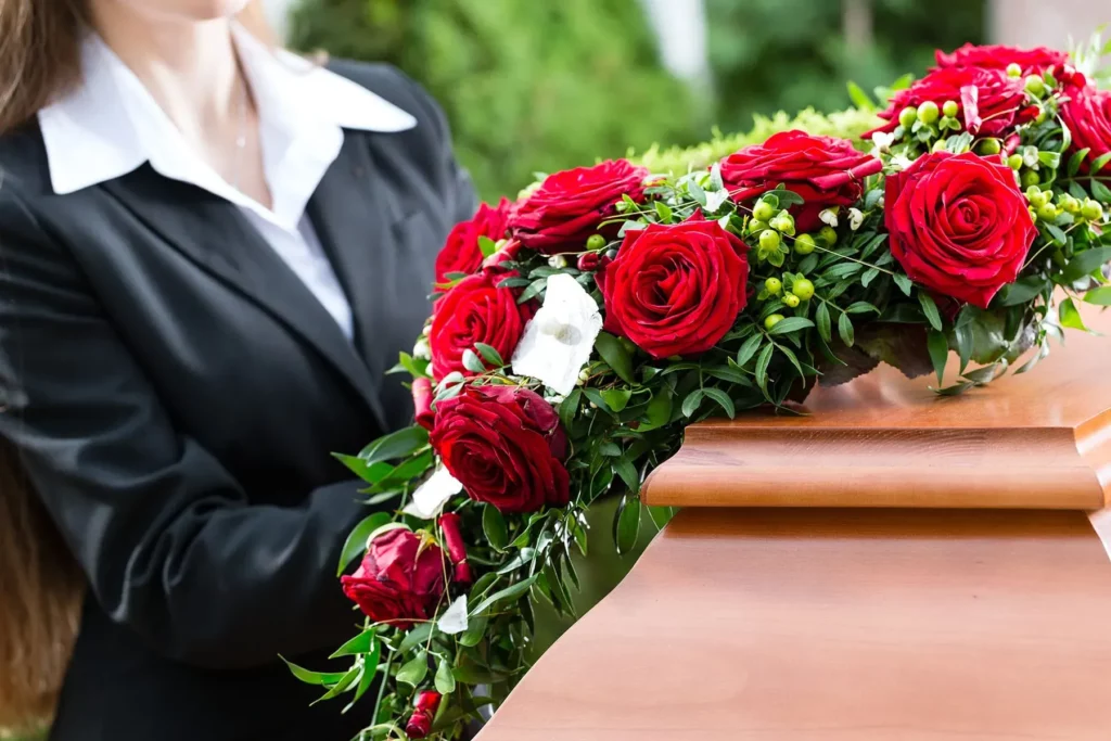 Femme en costume noir disposant une composition florale de roses rouges sur un cercueil en bois poli